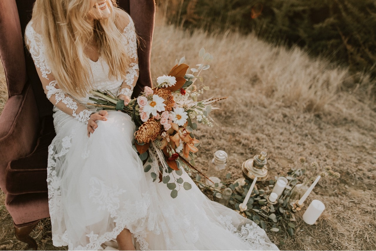 a bride in her long sleeve wedding gown, sitting in a field and holding her fall bouquet