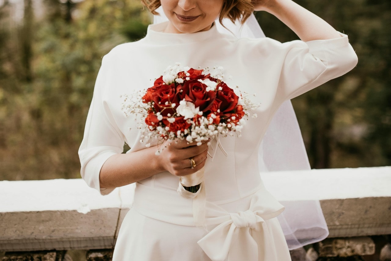 a bride wearing a dress with ¾ length sleeves and holding a bouquet with red roses