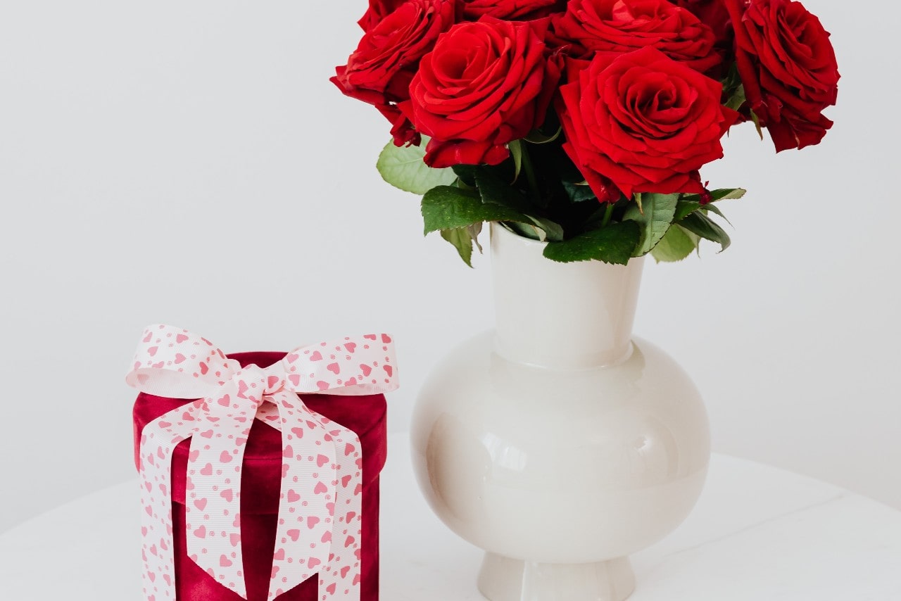 Velvet red cylinder box with a white and pink bow on top next to a boutique of red roses.