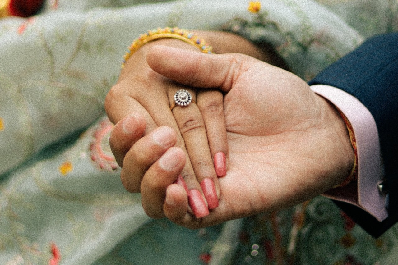 a man’s hand holding a woman’s, adorned with a halo engagement ring