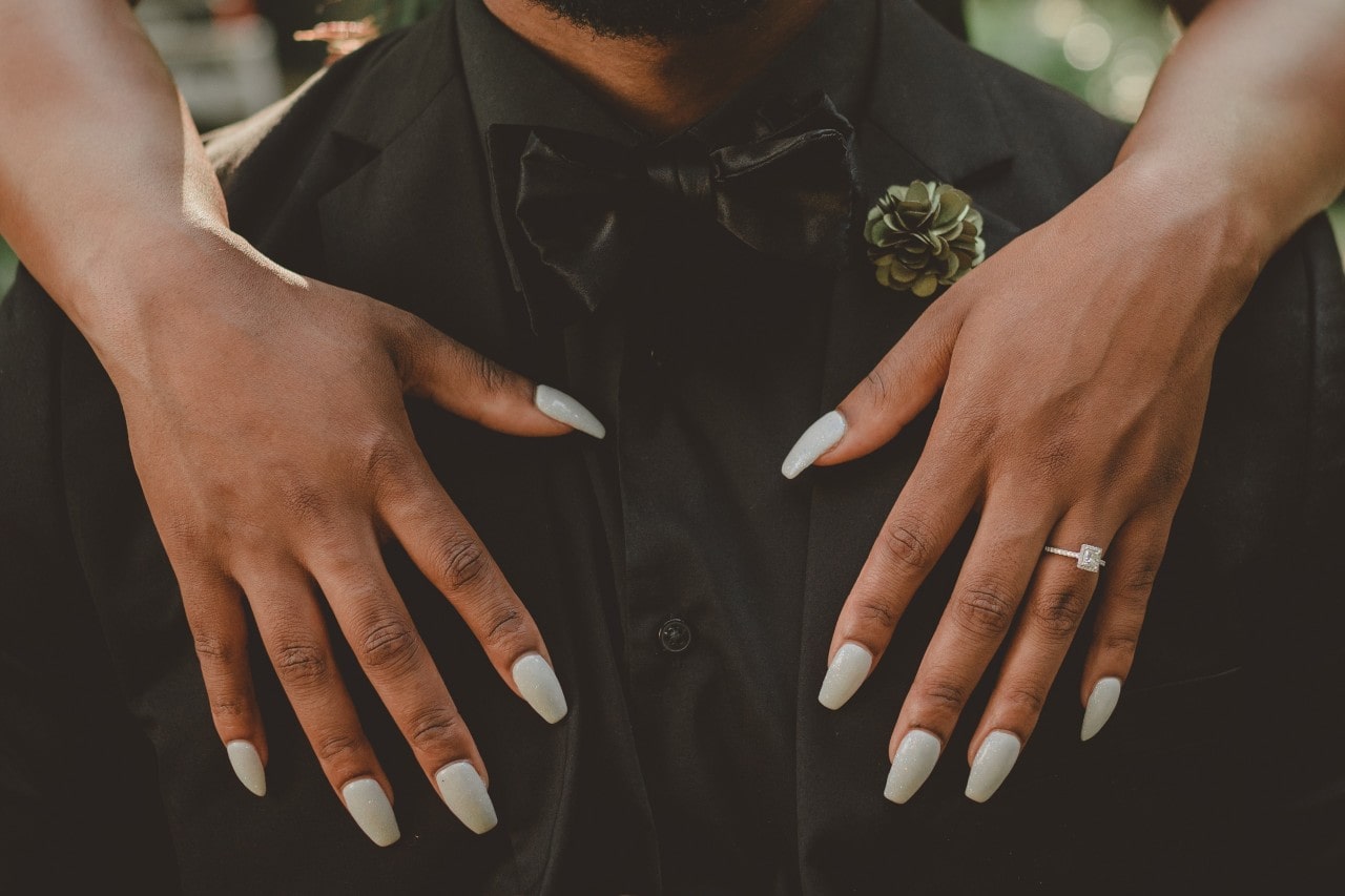 A close-up of a bride’s hands on her groom’s chest, a cushion-cut engagement ring on her finger.