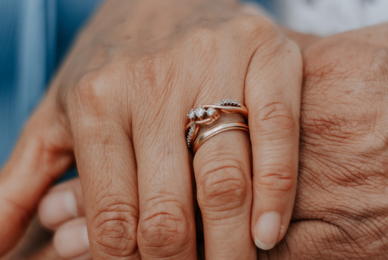 a woman holds a man’s hand, showing her gold wedding band and engagement ring.
