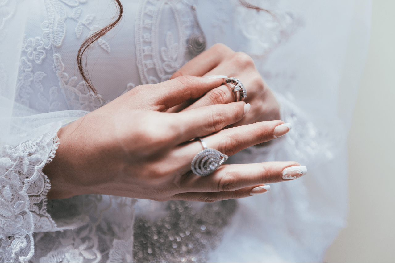 A close-up of a bride’s hands adorned with elegant silver rings, adjusting one of her rings against the backdrop of a lace wedding dress.