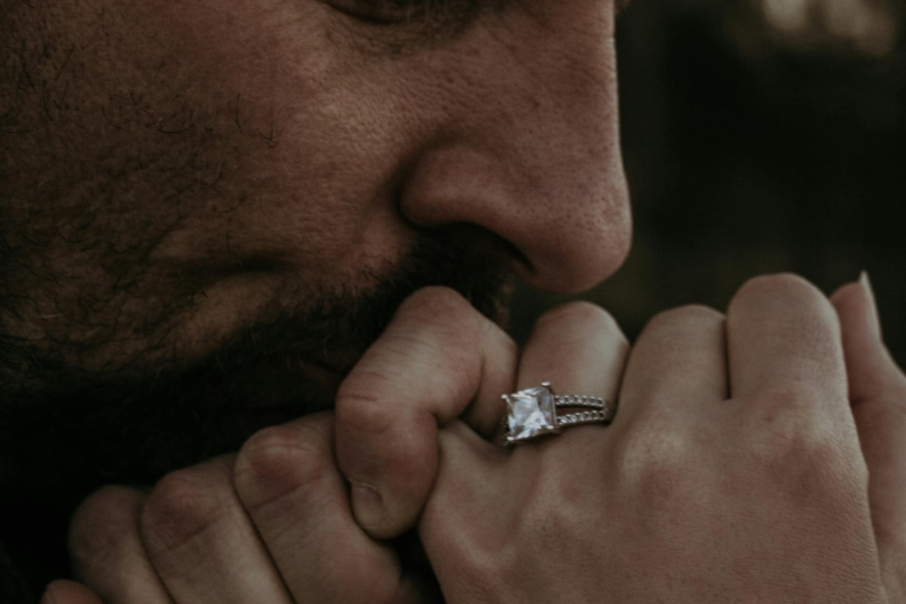 a man kissing a woman’s hand adorned with a princess cut engagement ring