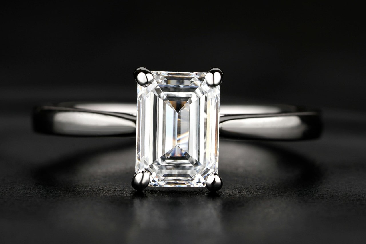 A close-up of a simple yet stunning emerald cut engagement ring on a black backdrop.